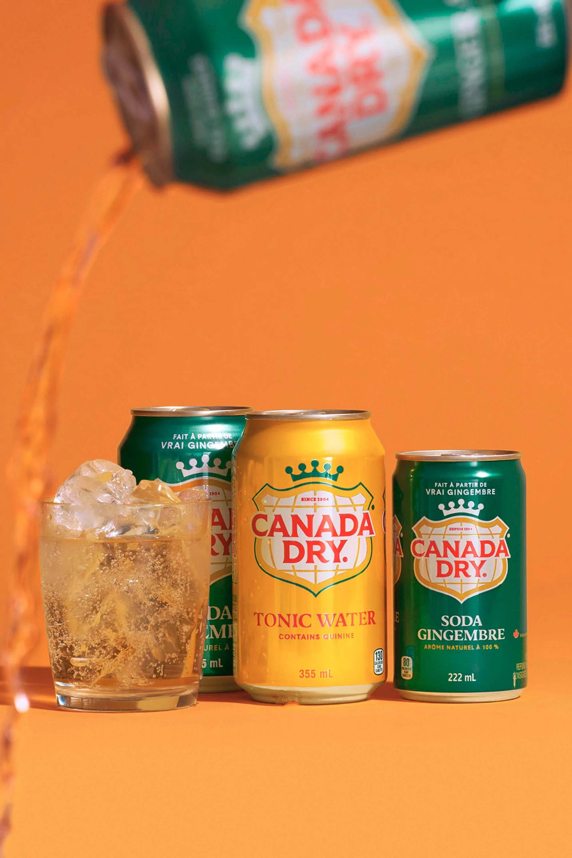 Several cans of Canada Dry and a glass with ice, arranged against an orange background, showcasing a refreshing and visually appealing beverage still life.