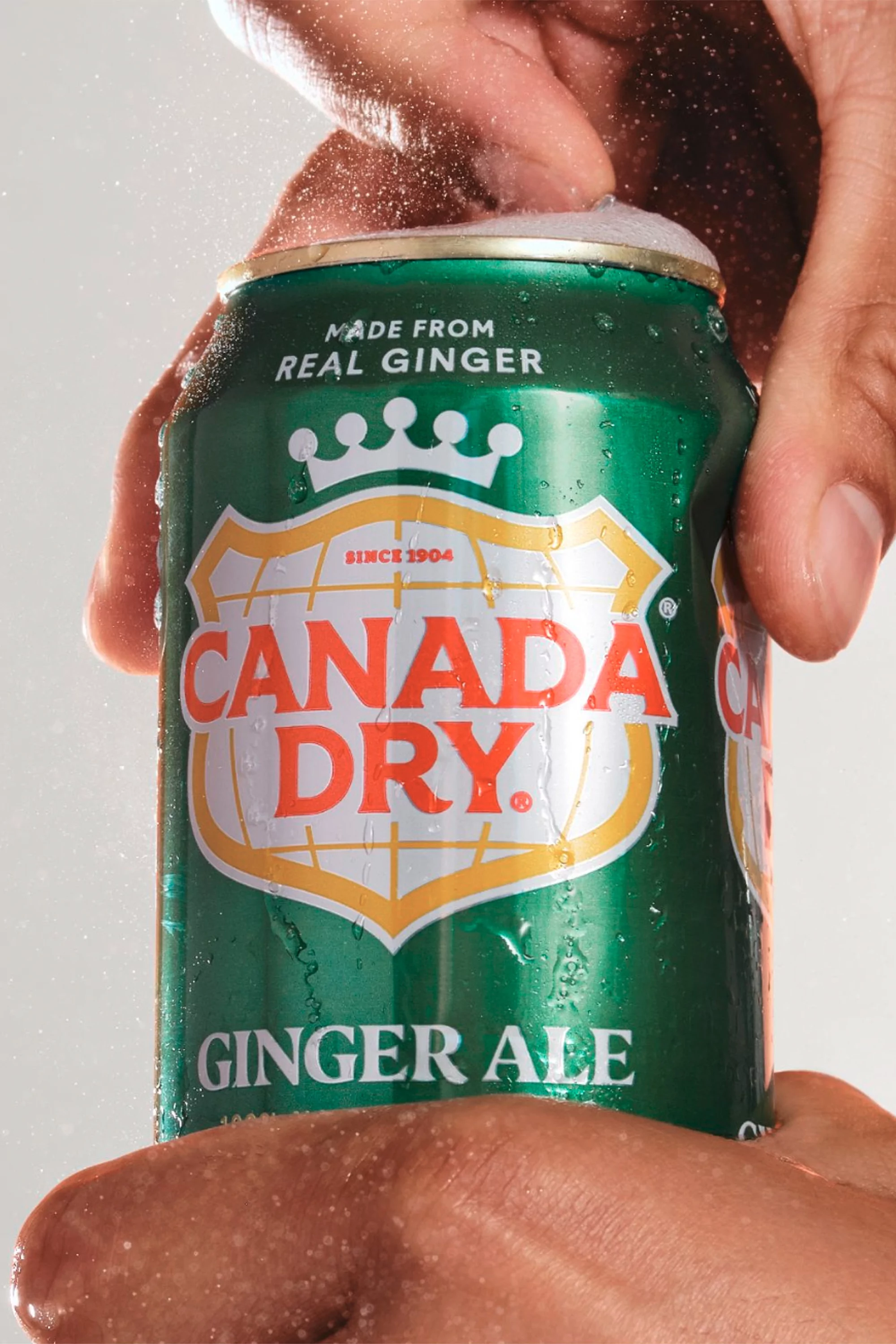 Close-up of a person's hands opening a chilled green can of Canada Dry ginger ale with condensation on the can, set against a white backdrop.