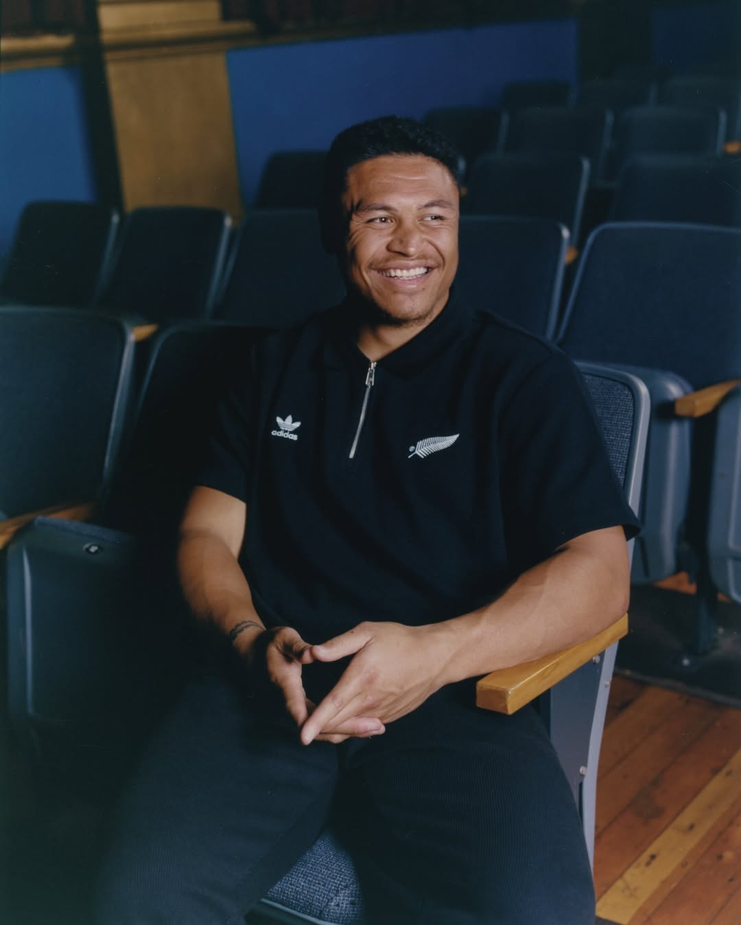 Smiling man in a black polo shirt and dark pants sits in an auditorium seat, hands clasped, with rows of empty seats behind him.