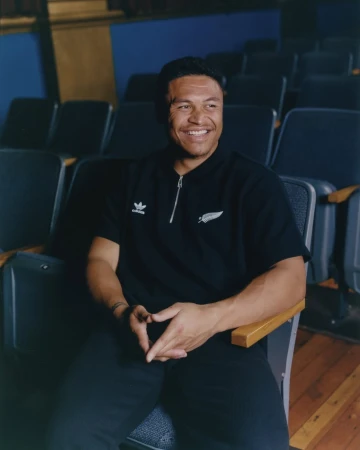 Smiling man in a black polo shirt and dark pants sits in an auditorium seat, hands clasped, with rows of empty seats behind him.
