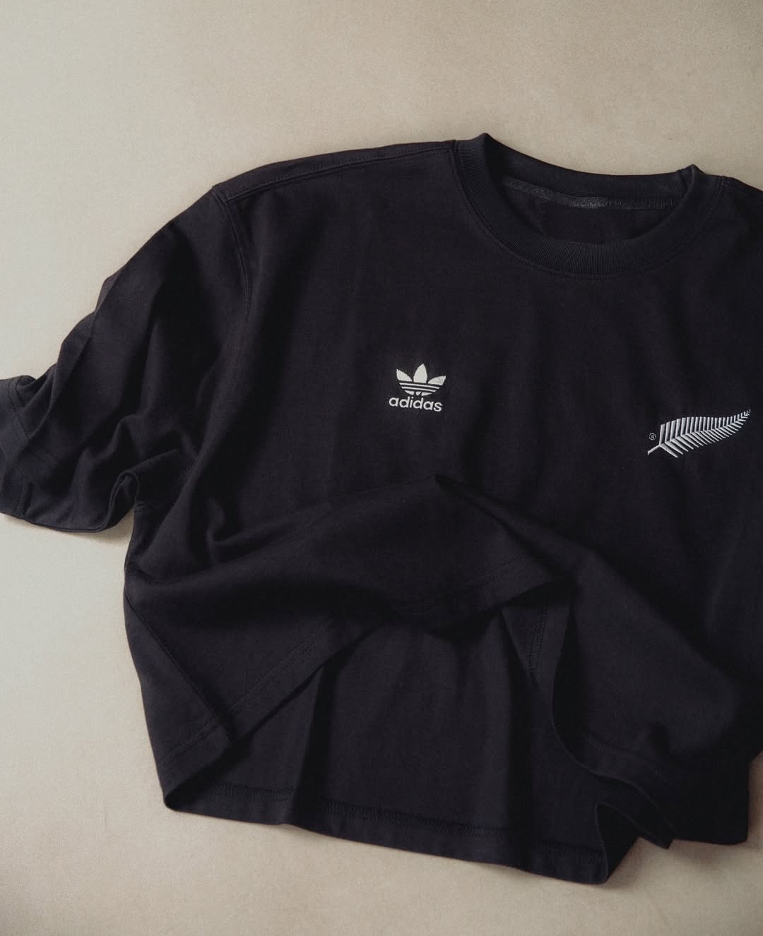 Black Adidas long sleeve shirt featuring the Adidas logo and fern design, laid on a light beige surface in a studio setting.