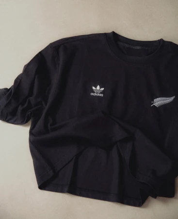 Black Adidas long sleeve shirt featuring the Adidas logo and fern design, laid on a light beige surface in a studio setting.