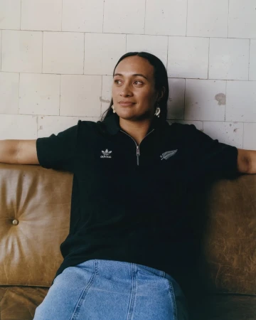 Woman with long dark hair wearing a black Adidas zip-up shirt and denim skirt is sitting on a brown couch against a tiled wall.