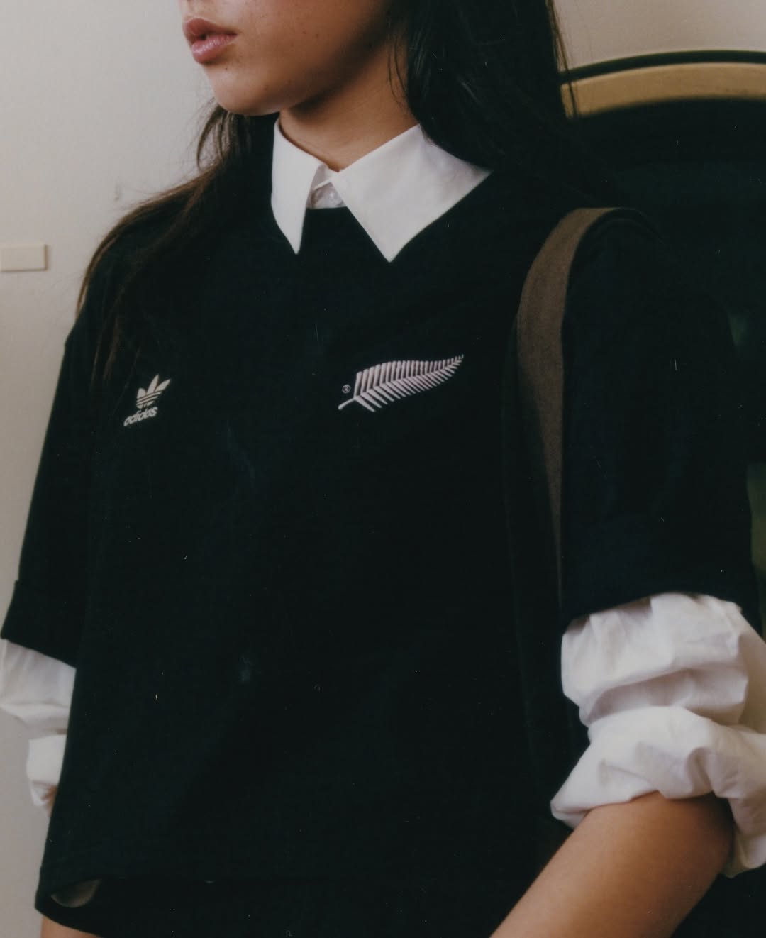 Woman in a black Adidas shirt with a white fern logo layered over a white collared shirt indoors, showing shoulders up.