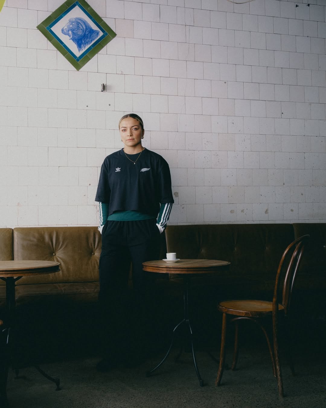 Young woman in a black Adidas athletic shirt stands in a cafe, looking at the camera. Behind her is a tiled wall and upholstered seating.