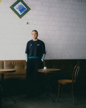 Young woman in a black Adidas athletic shirt stands in a cafe, looking at the camera. Behind her is a tiled wall and upholstered seating.
