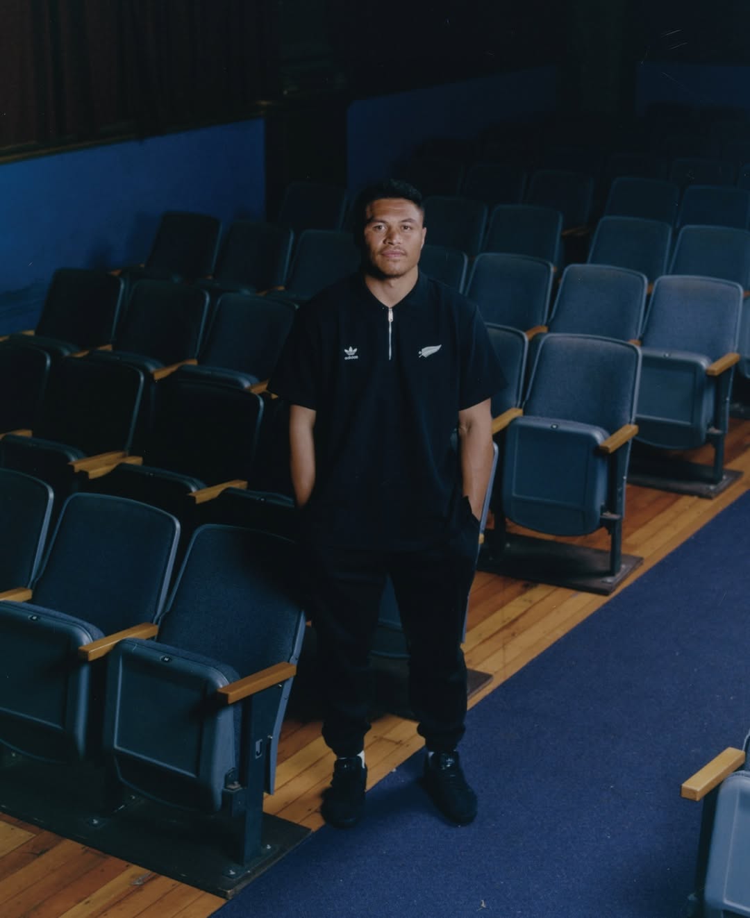 Adult male posing in a black polo shirt and black pants inside an empty theater with rows of dark blue seating and hardwood floors.