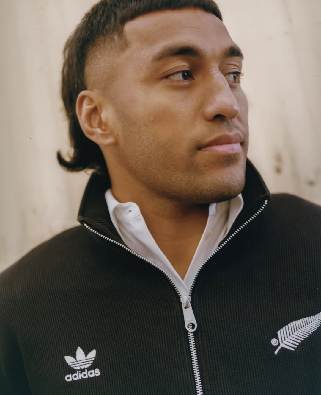Headshot of a stylish adult male with a modern mullet, wearing an Adidas track jacket, against a neutral background. Close-up view.