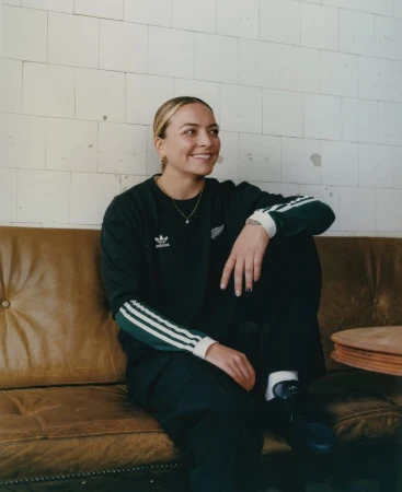 Smiling woman in Adidas tracksuit sits on a brown leather couch against a white-tiled wall in an indoor environment with soft lighting.