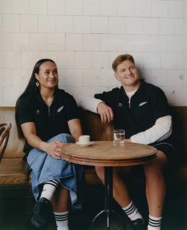 Two people sitting at a round wood table with a coffee cup and water glass, wearing matching black Adidas polo shirts in an indoor setting.