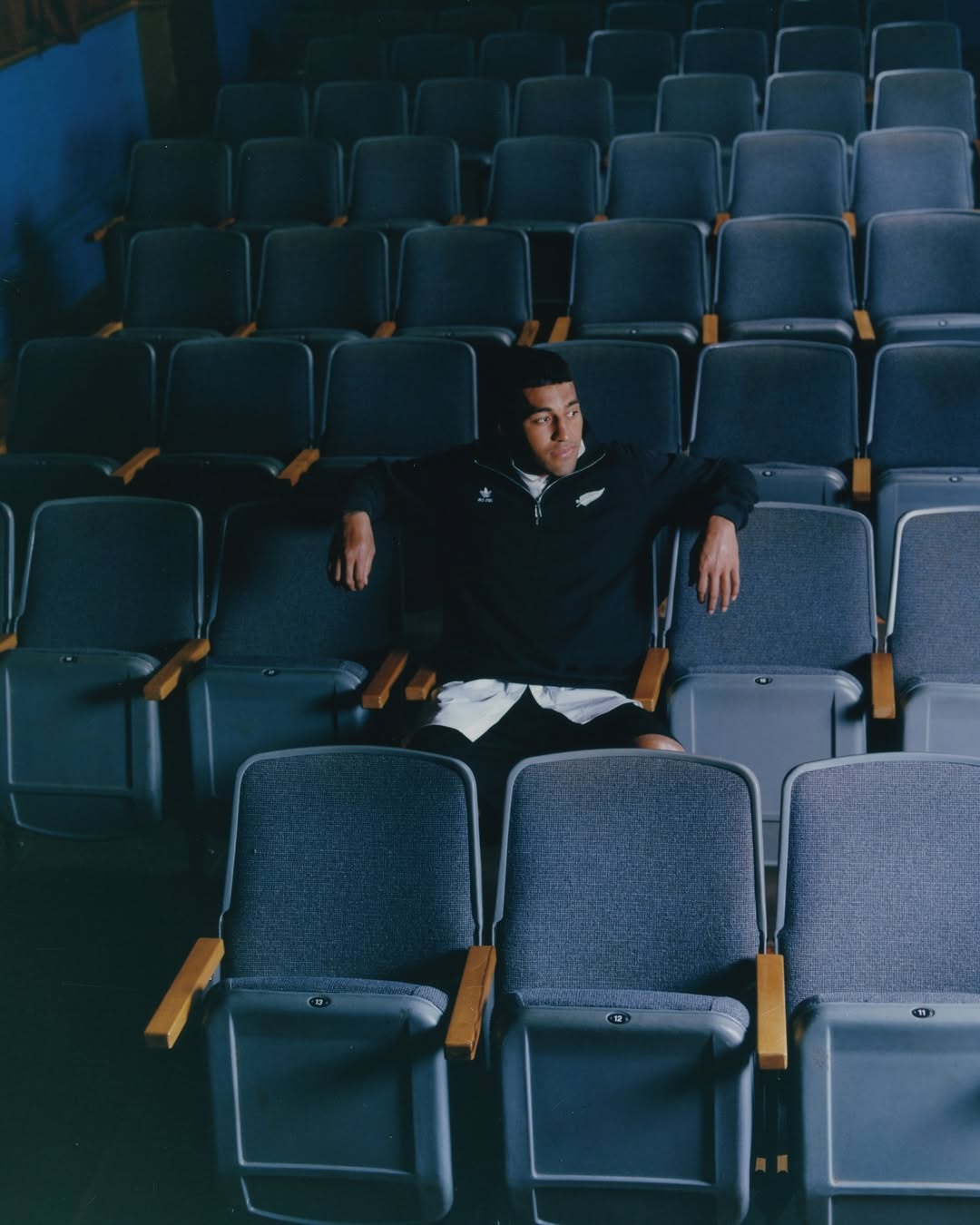 Man sits alone in a dimly lit theater, wearing a black top and white shorts, looking left, surrounded by rows of empty blue seats.