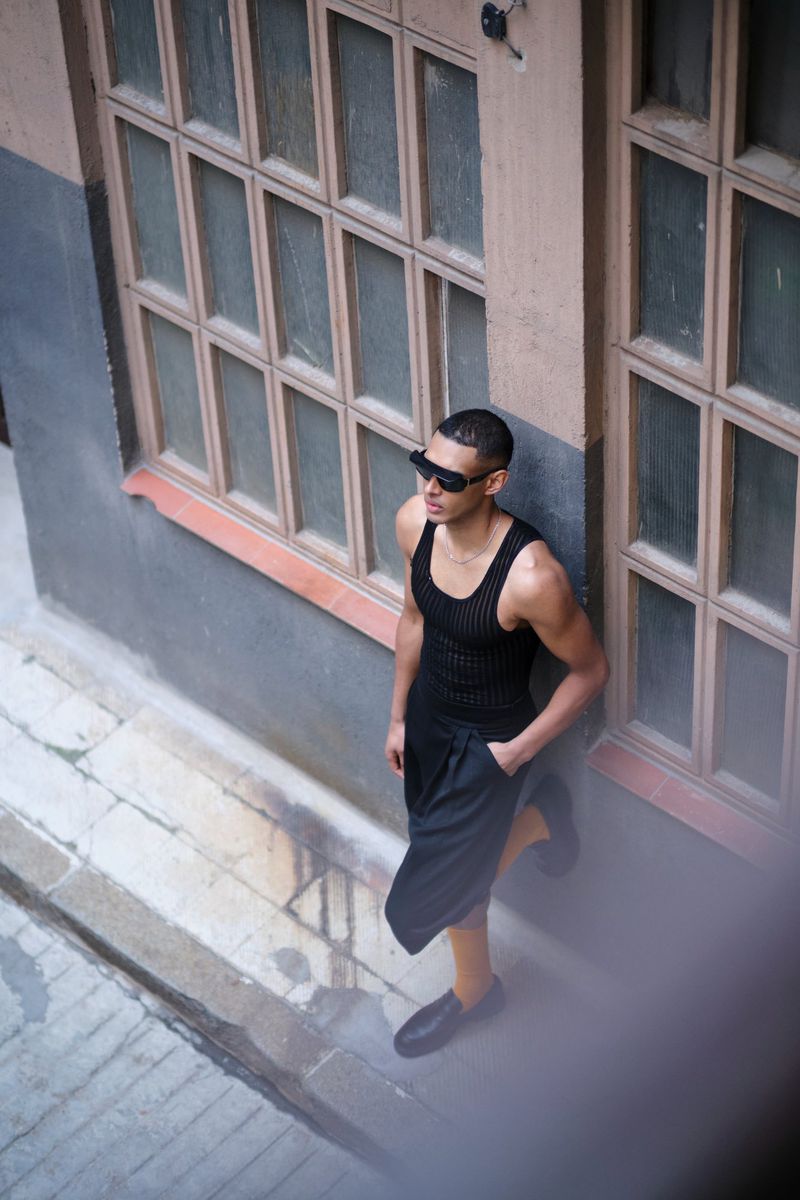 An adult male stands in an urban alleyway wearing a tank top and shorts, complemented by sunglasses and black shoes, under daylight.