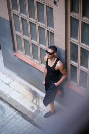 An adult male stands in an urban alleyway wearing a tank top and shorts, complemented by sunglasses and black shoes, under daylight.