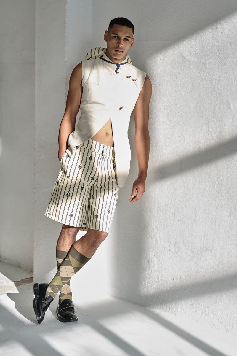 Man in sleeveless shirt and striped shorts poses against white wall with natural light. He wears patterned socks and black loafers.