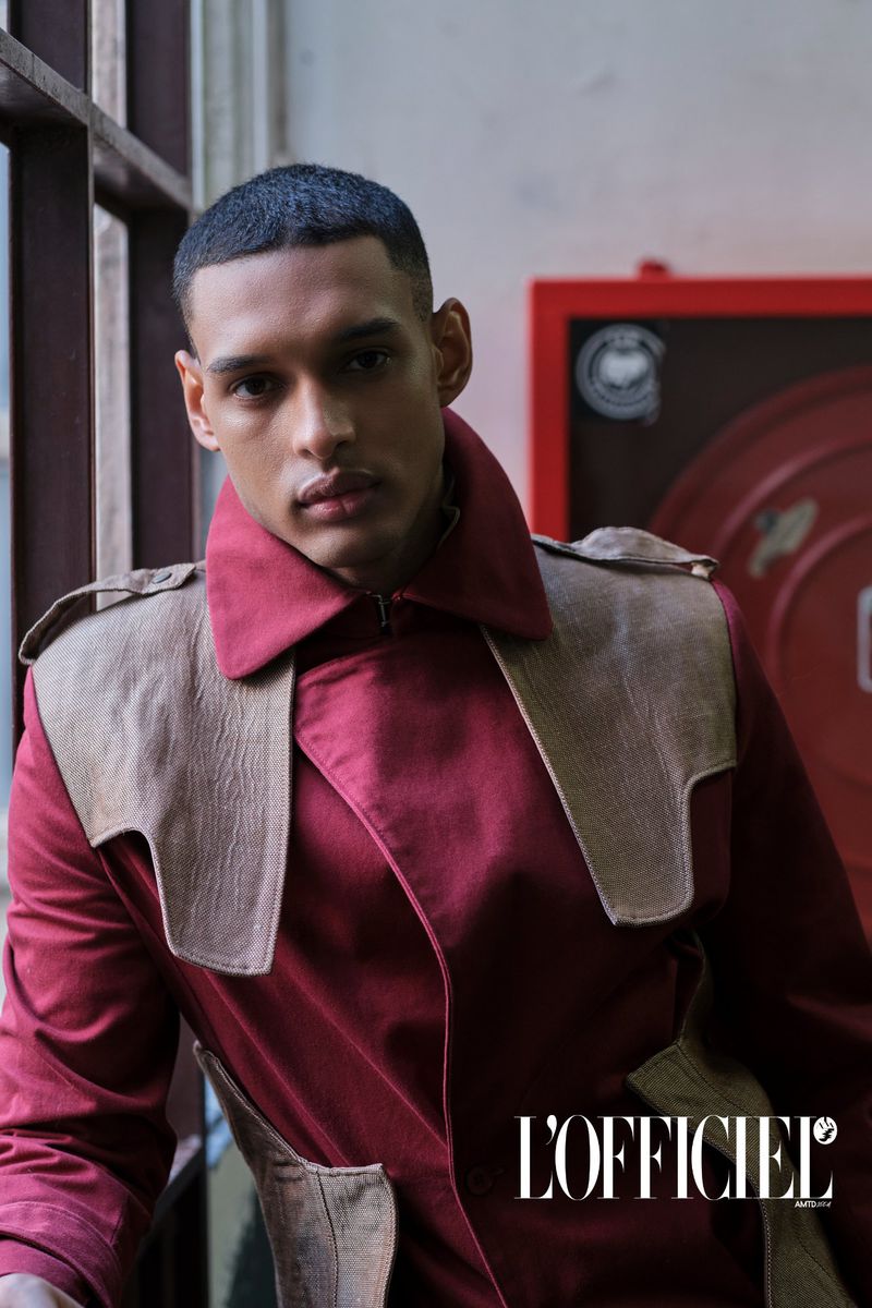 Editorial shot of a young adult male in a maroon jacket with tan panels, inside near a window, in front of a red industrial object.