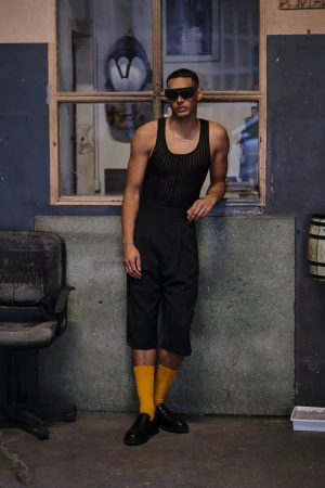 Man in tank top and shorts leans against a wall in a warehouse-style space wearing black sunglasses and yellow socks with black dress shoes.