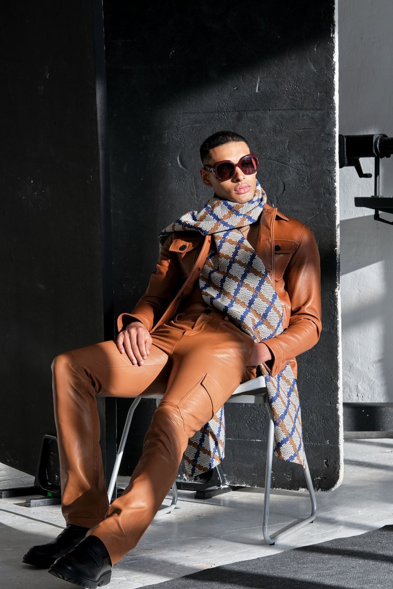 Stylish man in brown leather jacket and pants, posing in sunglasses and patterned scarf while seated, with studio lighting and black background.