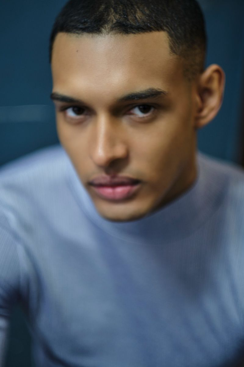 Close-up portrait of a young adult male of mixed ethnicity wearing a light blue turtleneck sweater, gazing directly at the camera.