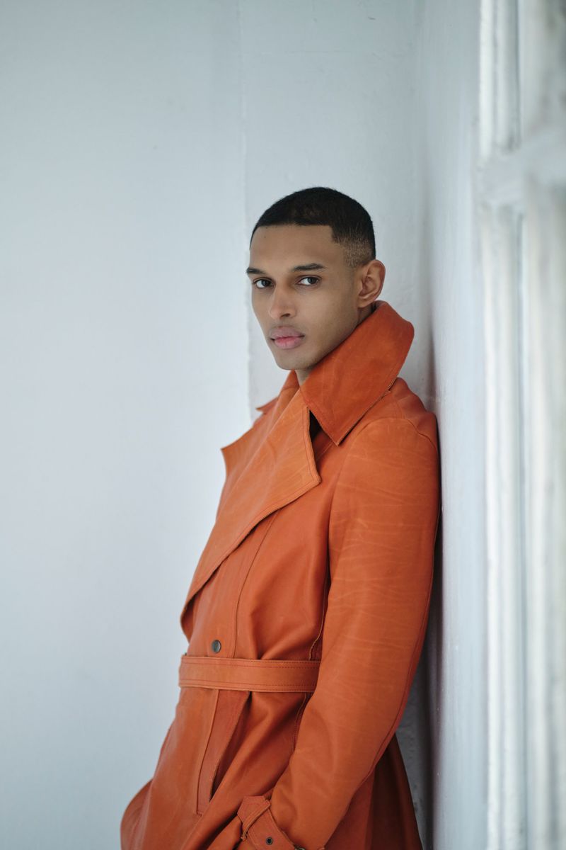 Portrait of a dark-skinned man in an orange trench coat leaning against a white wall with a window, gazing at the camera.