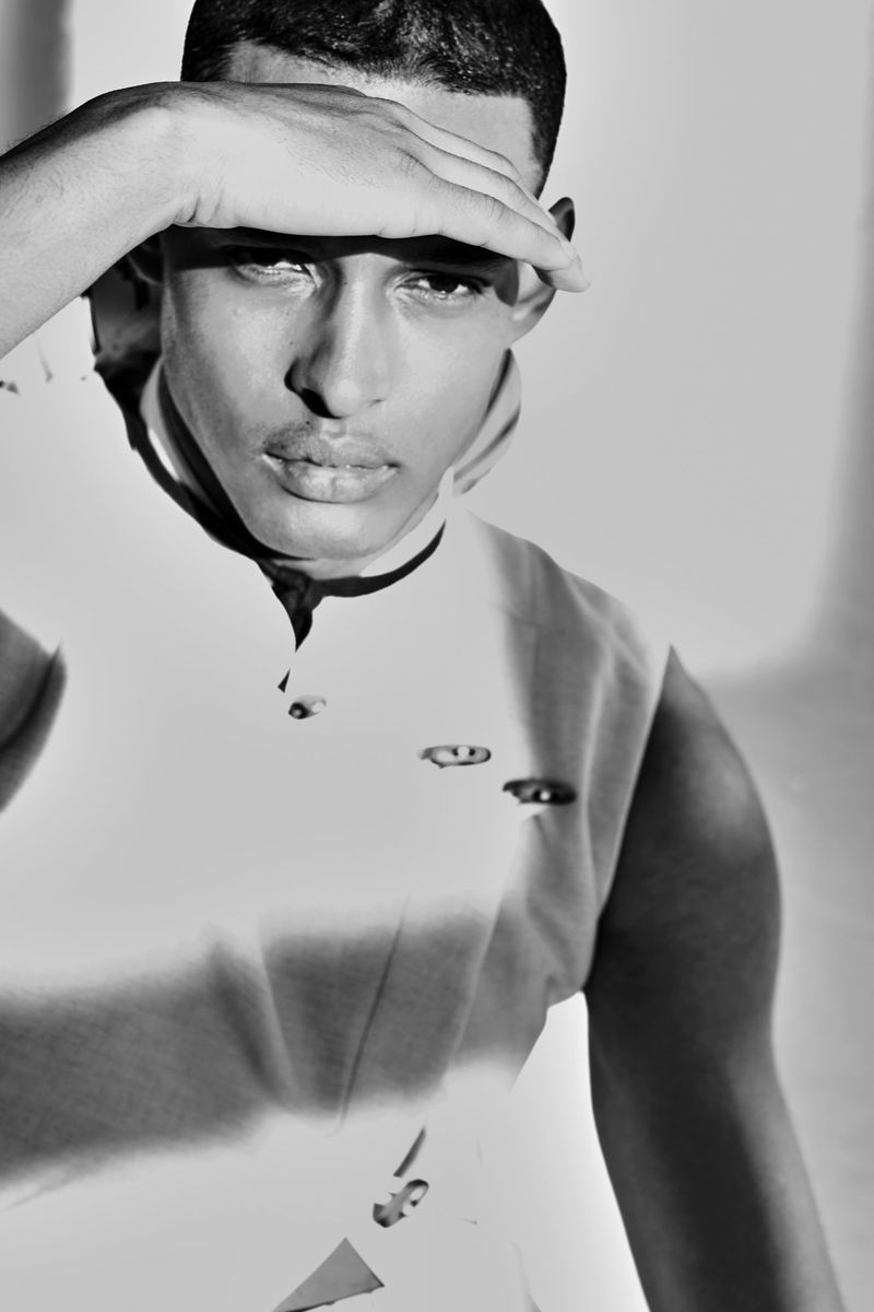 Portrait of a young adult man with dark hair looking at the viewer, shielding his eyes with his hand, wearing a light vest.