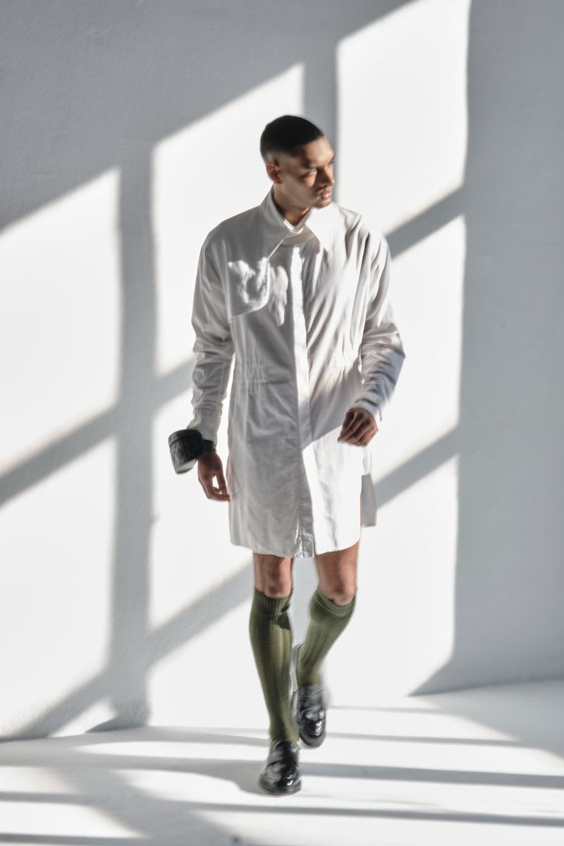 A slender male strides in a pristine white coat against a backdrop of geometric window light, complemented by olive socks and glossy shoes.