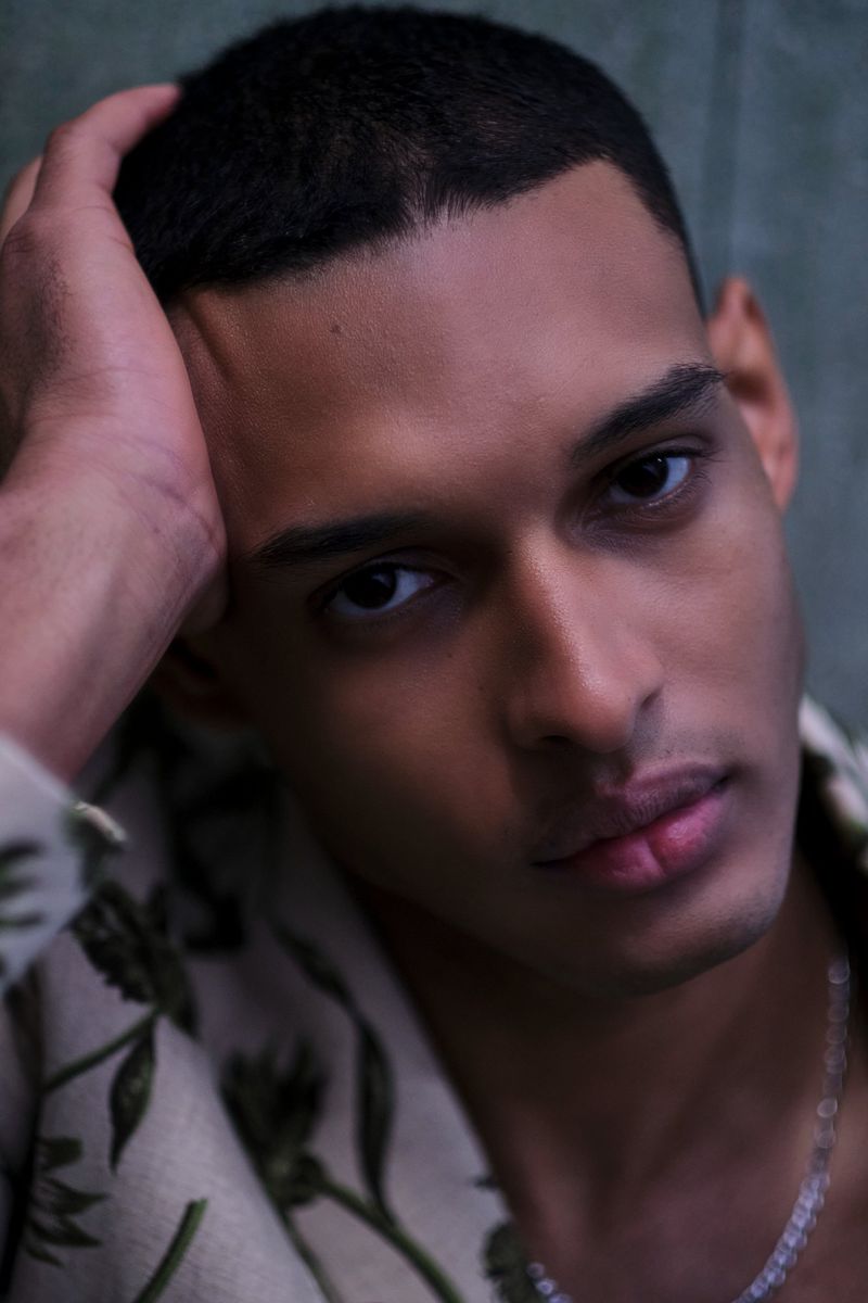 Portrait of a young adult man with dark hair, wearing a floral shirt and silver chain, indoors with neutral background, looking at camera.