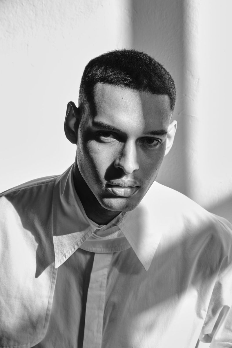 Black and white portrait of a handsome adult male with a serious expression, wearing a button-down shirt and standing in a light-filled room.