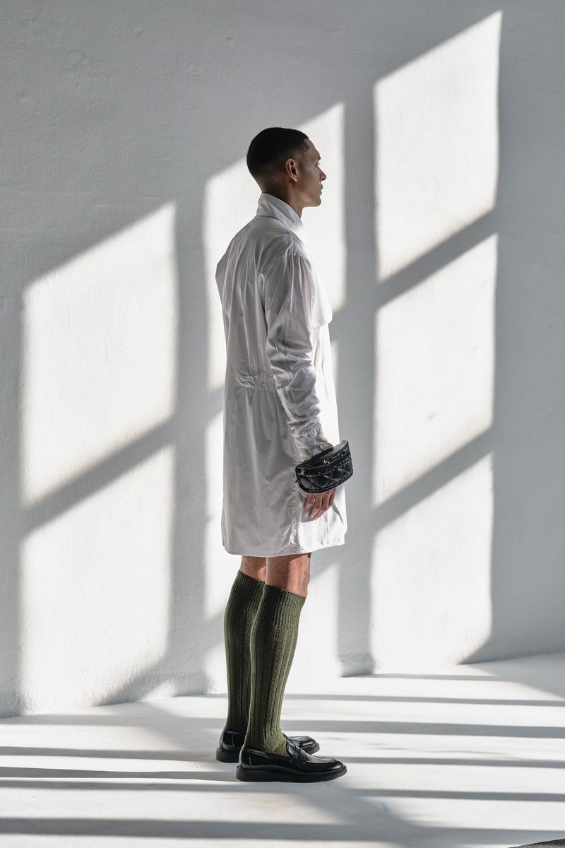 A slender Black male model in a white coat, knee socks, and loafers stands in a naturally lit white room, casting strong light and shadows.