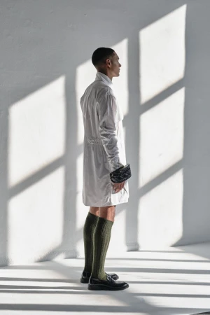 A slender Black male model in a white coat, knee socks, and loafers stands in a naturally lit white room, casting strong light and shadows.