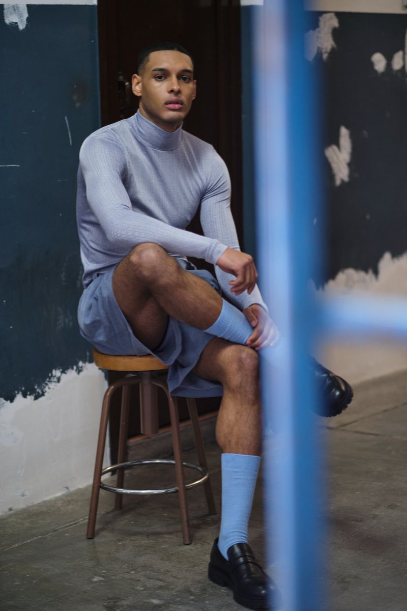 A young adult man sits on a stool in a studio, wearing a light blue turtleneck, matching shorts and socks, and black shoes, looking at the camera.