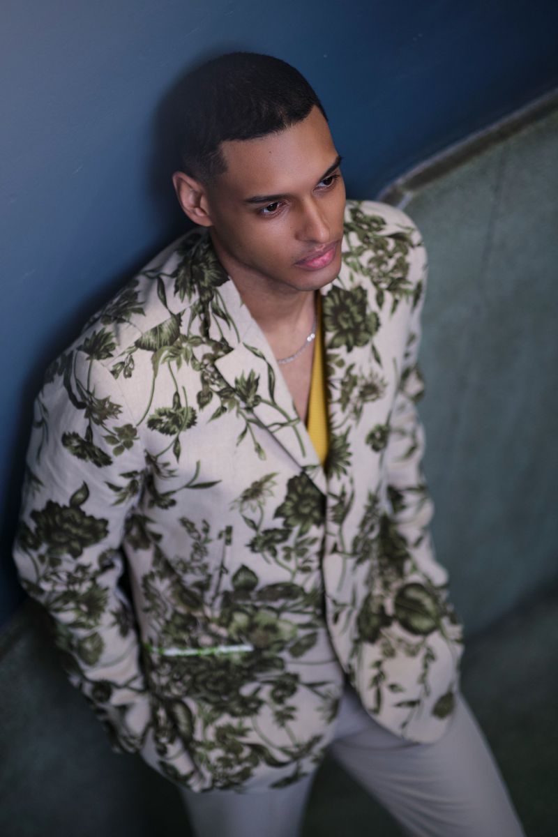 Stylish adult male poses indoors wearing floral blazer and light pants, looking off-camera with serious expression in natural light.