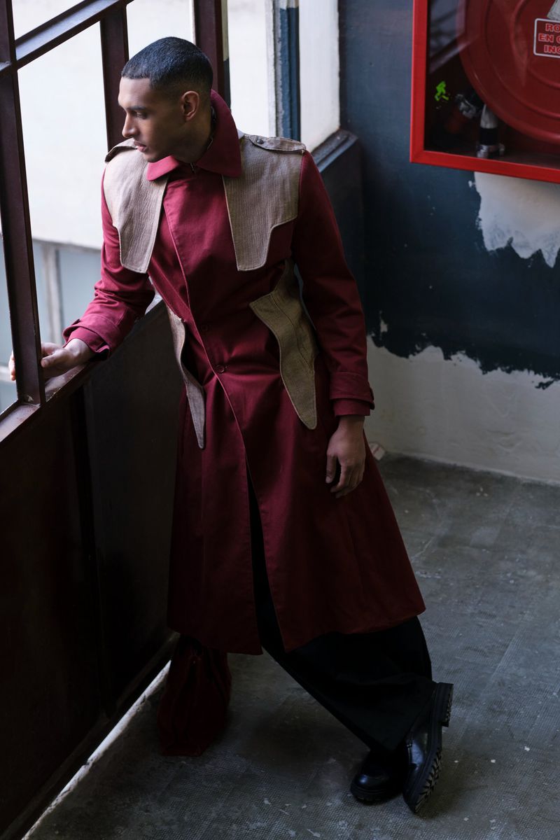 Man in burgundy coat and black pants poses on a staircase near a window and fire extinguisher for a fashion editorial shoot.