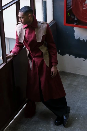 Man in burgundy coat and black pants poses on a staircase near a window and fire extinguisher for a fashion editorial shoot.