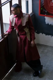 Man wearing a long red coat with beige panels looking out a window indoors