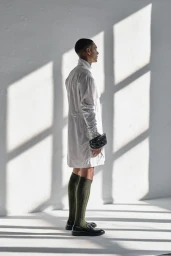Man wearing a white long shirt and green knee-high socks stands against a white wall with strong window shadows.