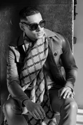 Man wearing sunglasses, leather jacket, and patterned scarf seated outdoors against a textured concrete wall.