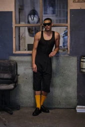 Man wearing dark sunglasses, black ribbed tank top, black shorts, and yellow socks leaning against a wall