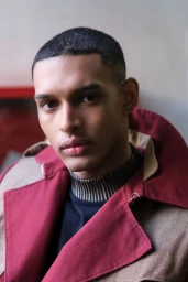 Close-up portrait of a young Black man wearing a burgundy coat and striped turtleneck for L'Officiel Baltics Essence project.