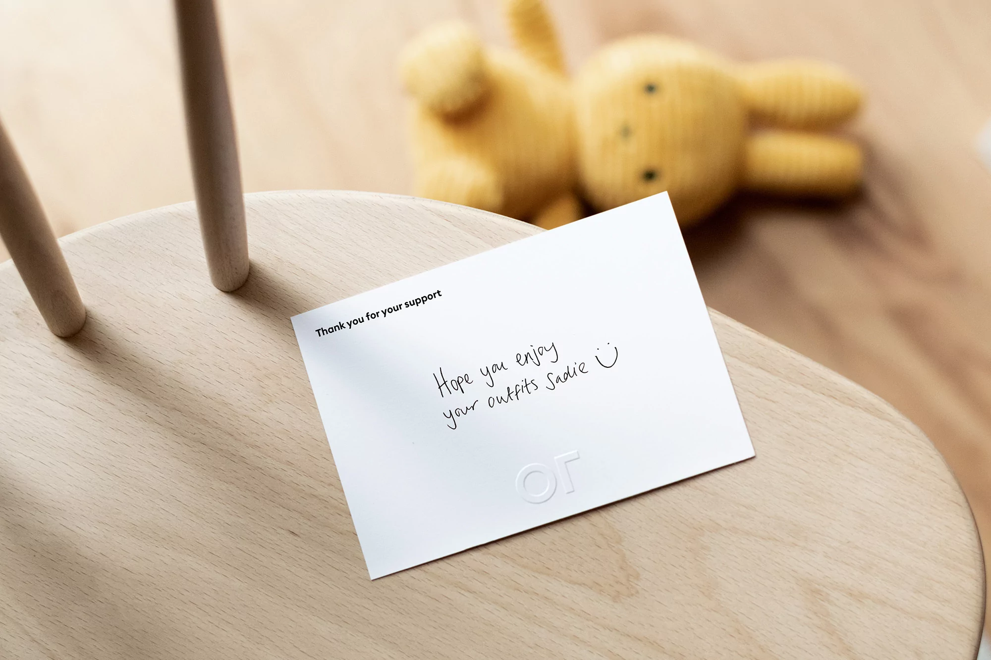 Close-up of a white card with a handwritten message resting on a light wooden chair, with a yellow stuffed bunny in the background.