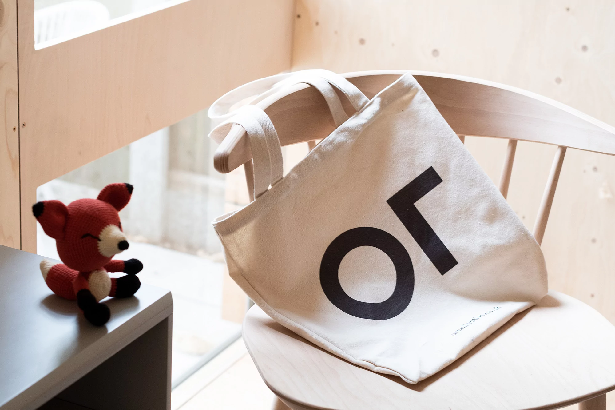 Crocheted fox toy and a beige tote bag with large black lettering sit on a light-colored wooden chair near a window.
