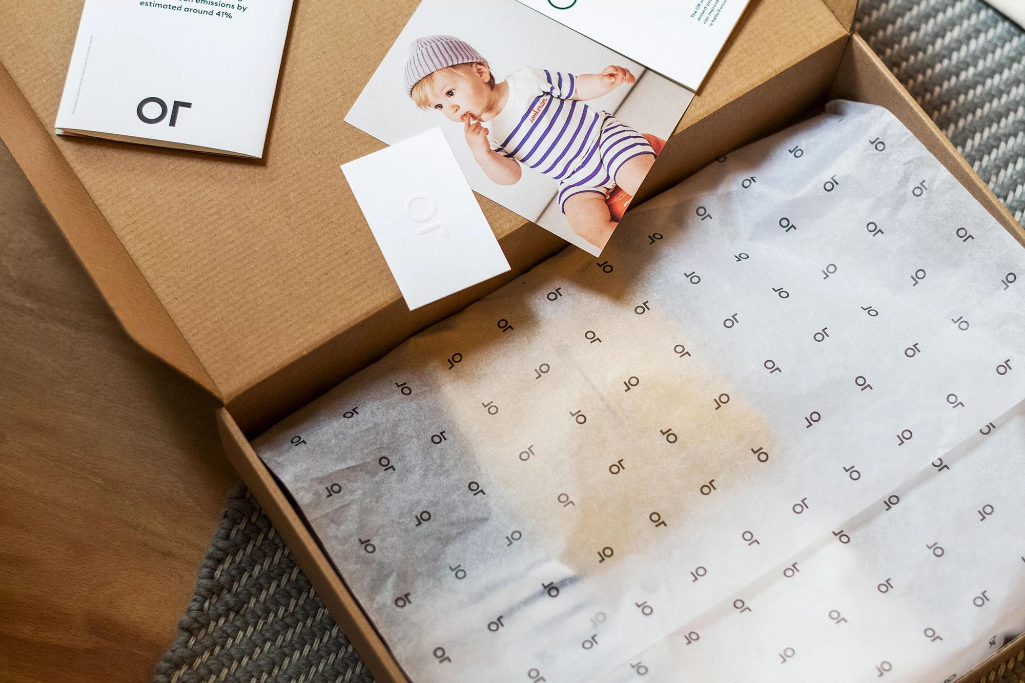 Open cardboard box containing a baby outfit, tissue paper, and brand cards on a wooden floor with a gray woven rug.