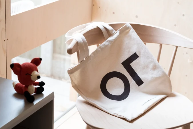 Cream canvas tote bag with black typography resting on a wooden chair next to a small crocheted fox toy.