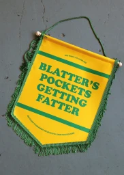 A yellow and green pennant hanging on a grey concrete surface, featuring bold text reading BLATTER S POCKETS GETTING FATTER.