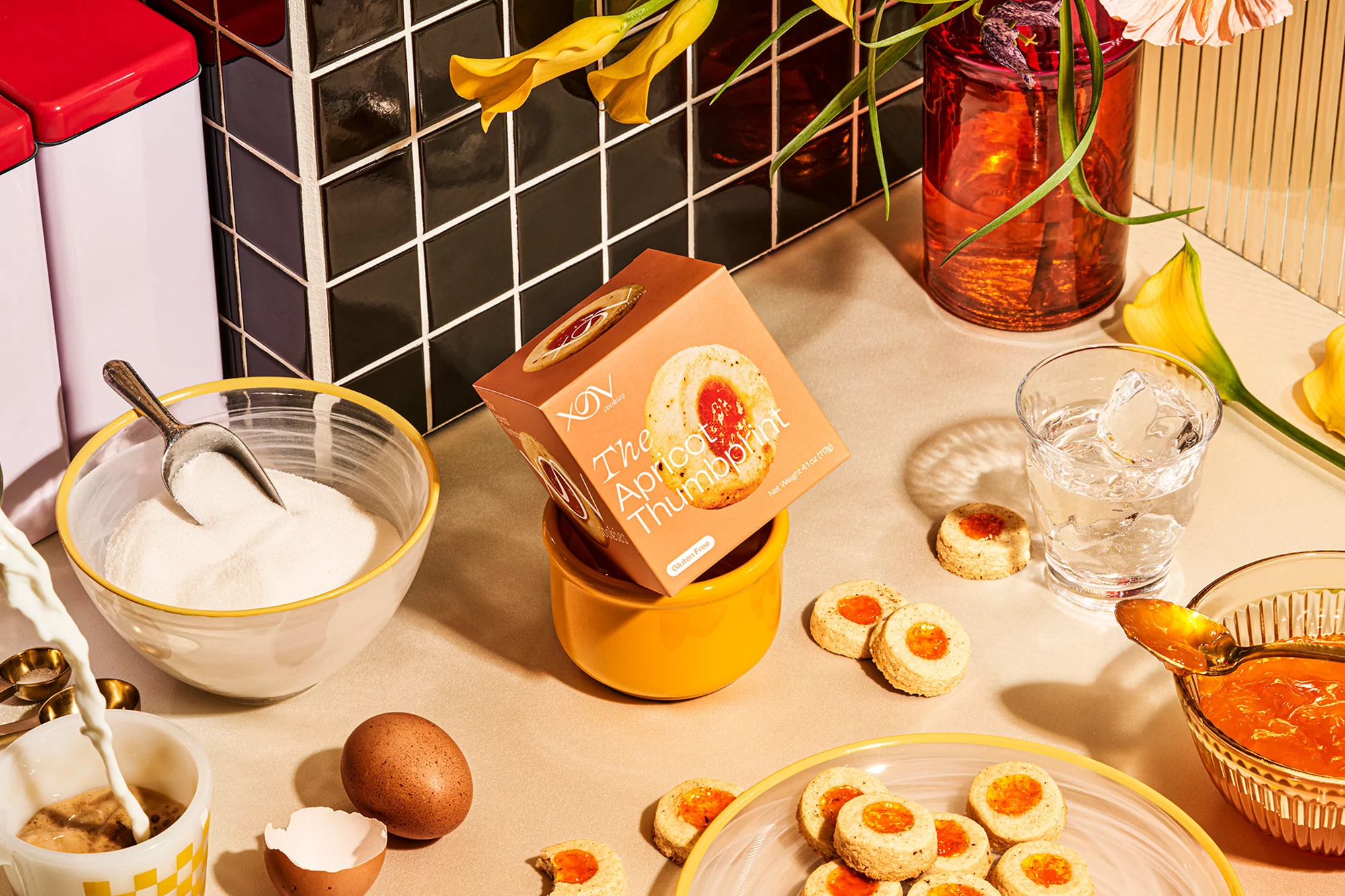 Thumbprint cookies presented on a table with sugar, eggs, apricot preserves, flowers, and a glass of water, set against a tiled wall.