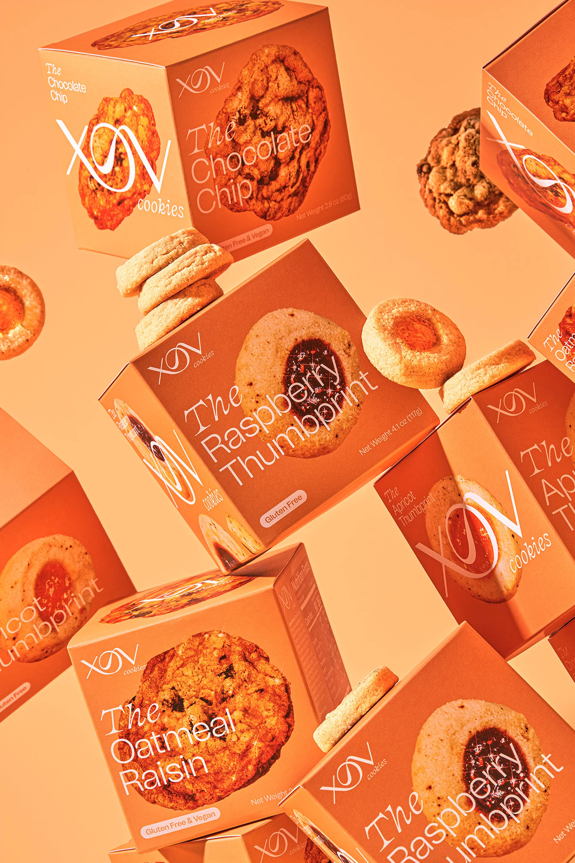 Still life shot of cookie boxes featuring chocolate chip, oatmeal raisin, raspberry thumbprint, and apricot thumbprint flavors against a peach backdrop.