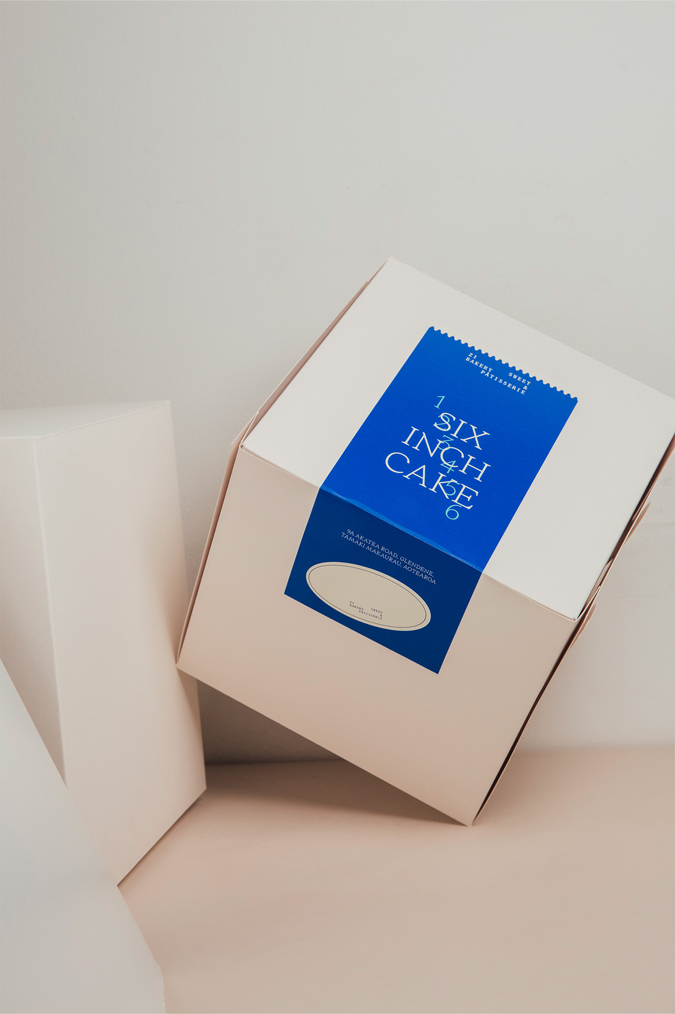 Cake box on beige surface. Square white cake box with blue label and white text. Gift and bakery packaging concept.