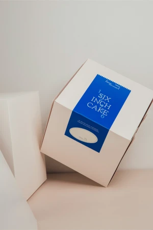 Cake box on beige surface. Square white cake box with blue label and white text. Gift and bakery packaging concept.