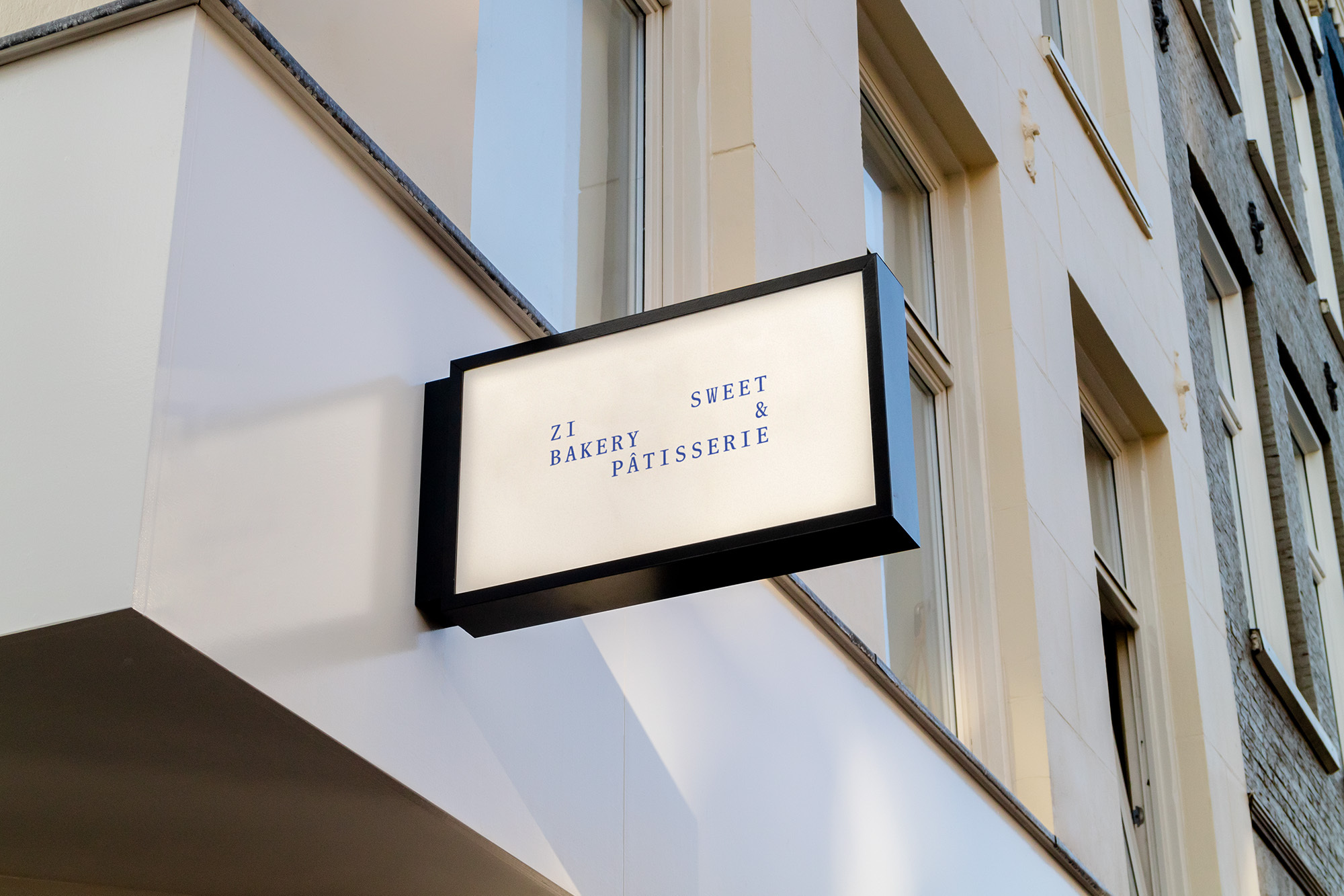 Exterior building shot featuring a lit sign for Zi Bakery & Pâtisserie with blue lettering in an urban setting with windows.