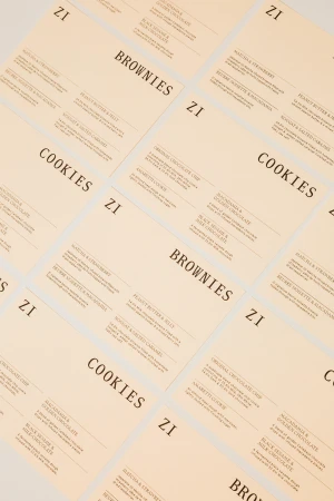 Arrangement of printed sheets displaying menus for cookies and brownies with product descriptions and brand logo on a neutral surface.