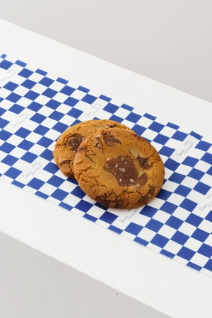 Two round chocolate chip cookies resting on blue and white checkered paper against a clean white background